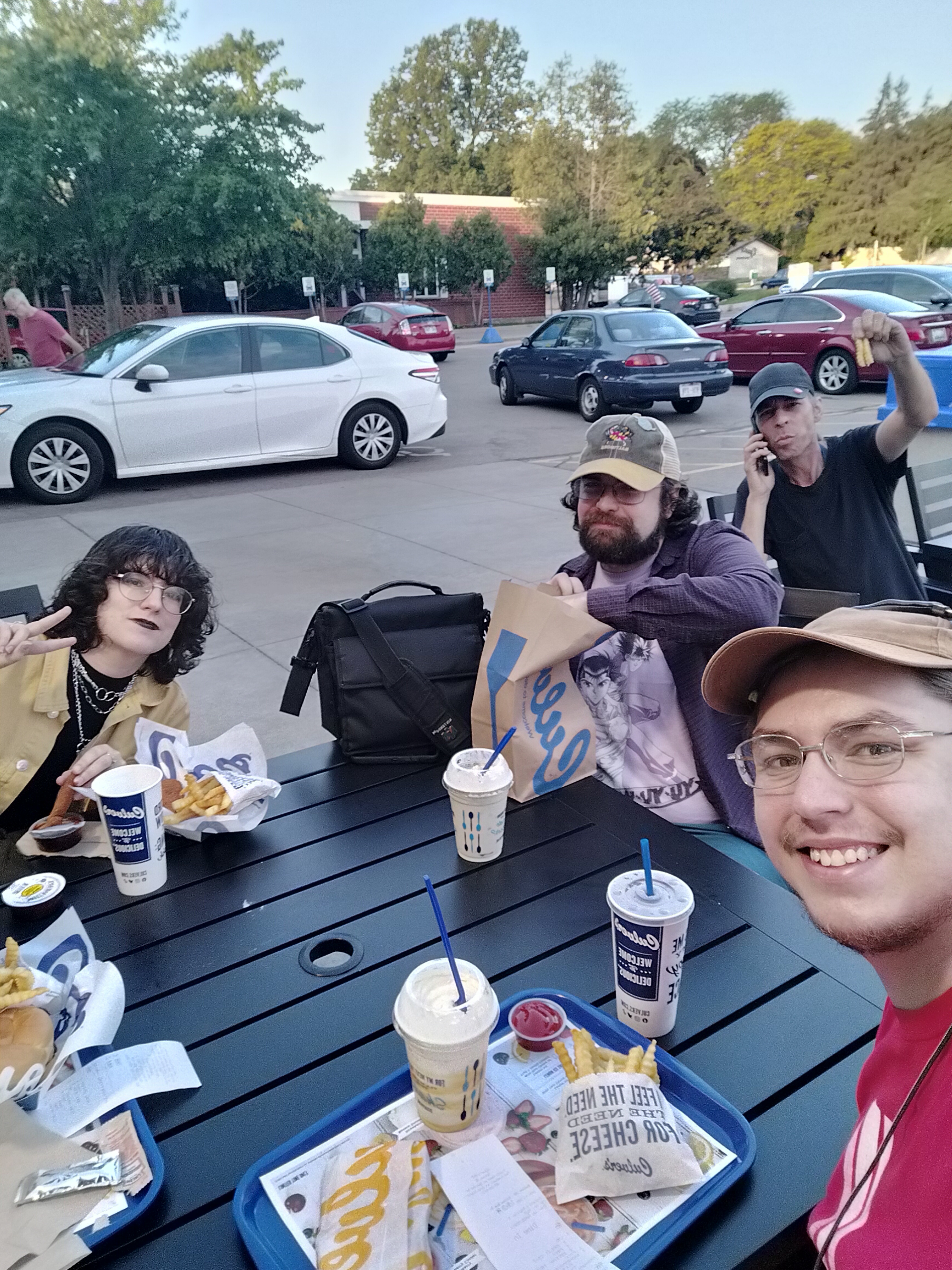 Five people (with one person off-camera) sitting at a table outside of a Culver's with five orders of food. Josiah, Christos, and Rob are sitting to the right.