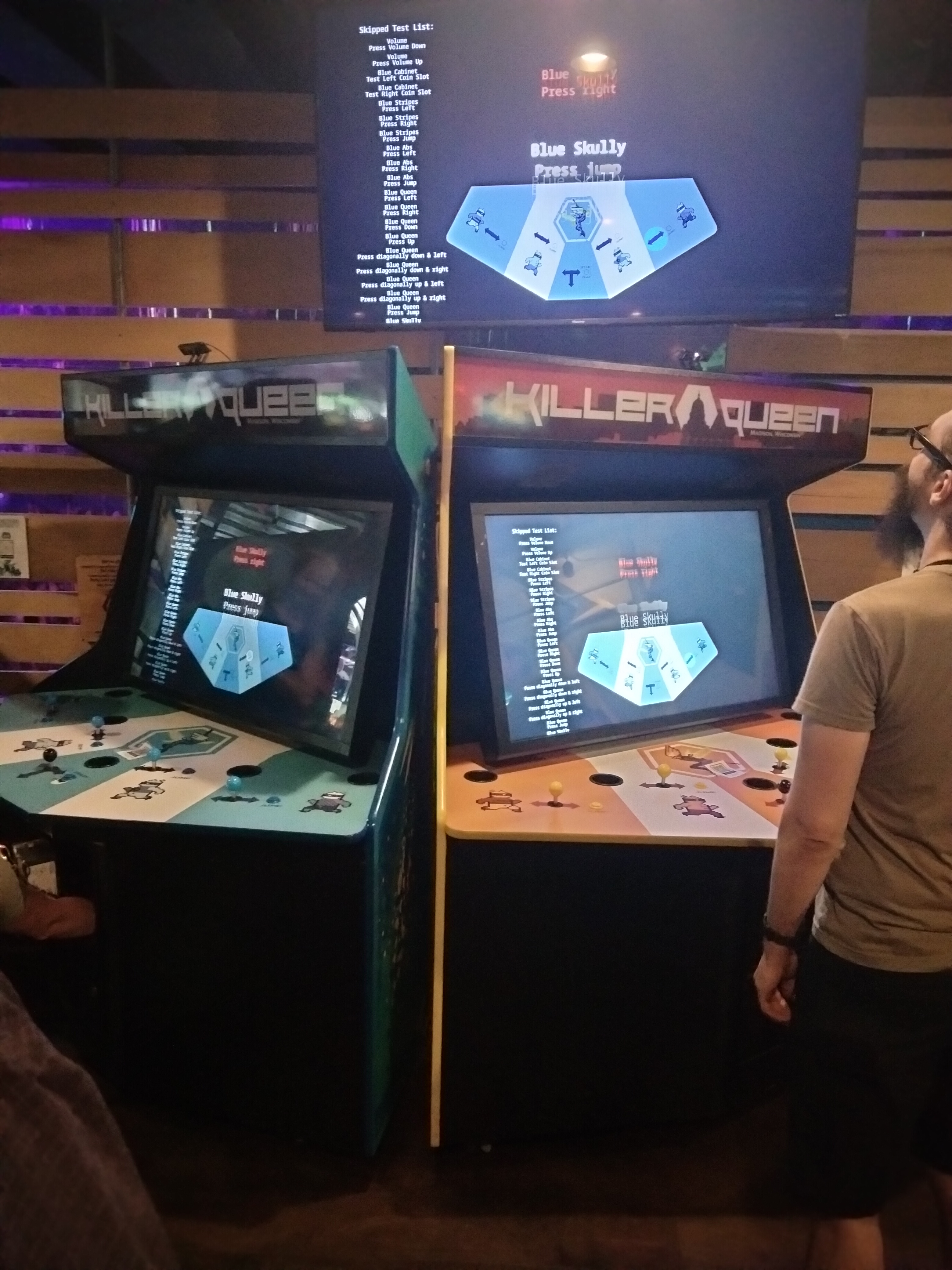 Two large arcade cabinets for Killer Queen, in blue and gold. A separate TV screen is above them, showing the same image that's on both cabinets. Some diagnostics tests are being run on the cabinets.